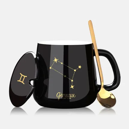 Black mug with Gemini zodiac constellation design, gold spoon, and lid on a light gray background