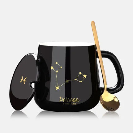 Black mug with gold constellation design, spoon, and lid on a light gray background