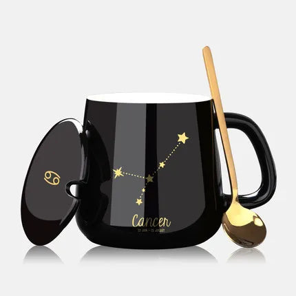 Black mug with Cancer zodiac design, gold spoon, and lid on a light gray background