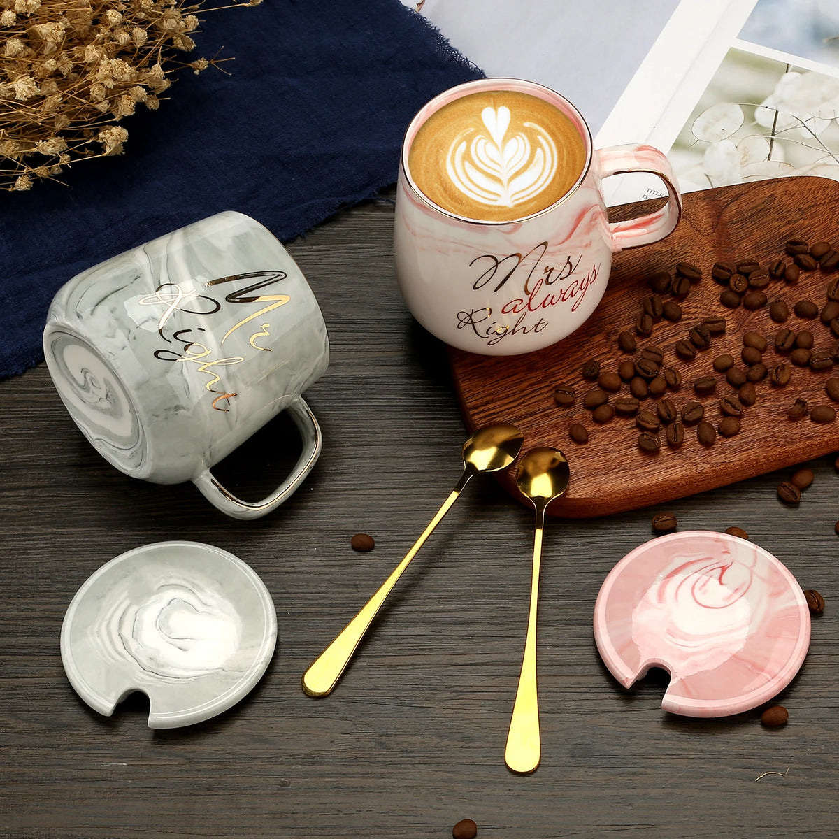 Two mugs with decorative text, a wooden board with coffee beans, and two gold spoons on a wooden surface.