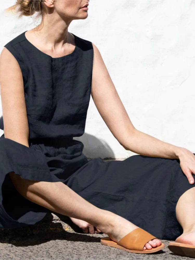 Woman wearing a black sleeveless dress sitting on a stone surface with a white wall in the background.