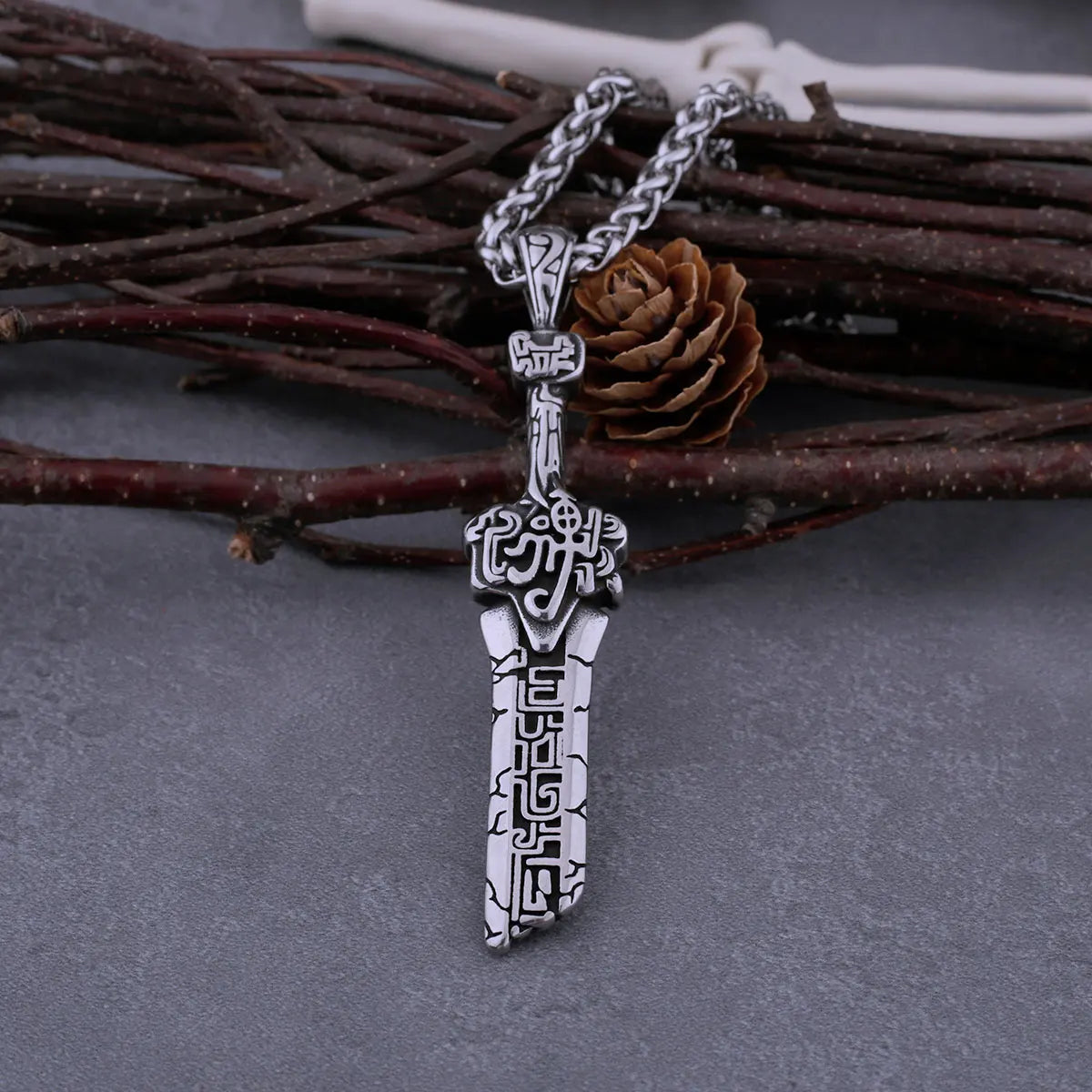 Silver dagger-shaped pendant on a chain against a textured gray background with twigs.