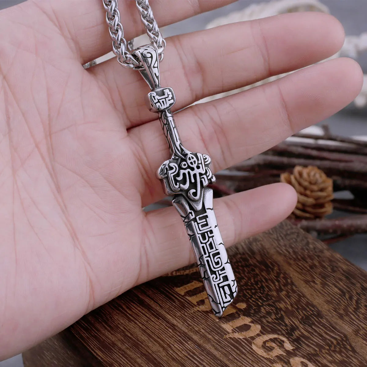 Silver dagger-shaped pendant on a chain held in a hand with a wooden surface in the background.